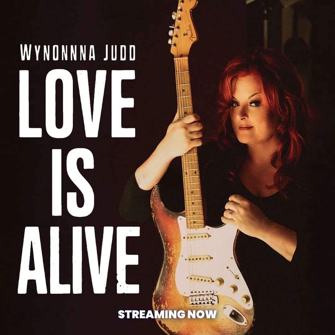 Wynonna Judd stands holding an electric guitar next to bold text reading "Love Is Alive" with "Streaming Now" at the bottom.